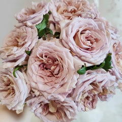 A close-up of a bouquet of pale blush pink roses with green leaves, photographed against a soft light background.