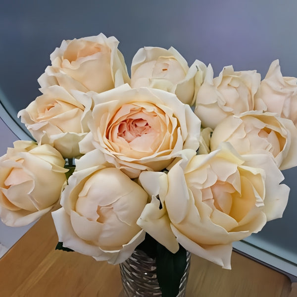 A bouquet of pale cream peony roses in a textured glass vase, set on a wooden surface against a gray background.